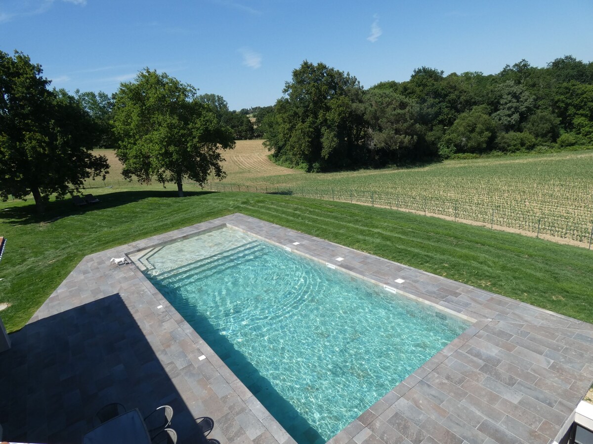 A rectangular swimming pool is set within a stone patio, reflecting clear blue water under a sunny sky. Lush green grass surrounds the pool, leading to a view of fields and trees in the background, creating a serene outdoor space.