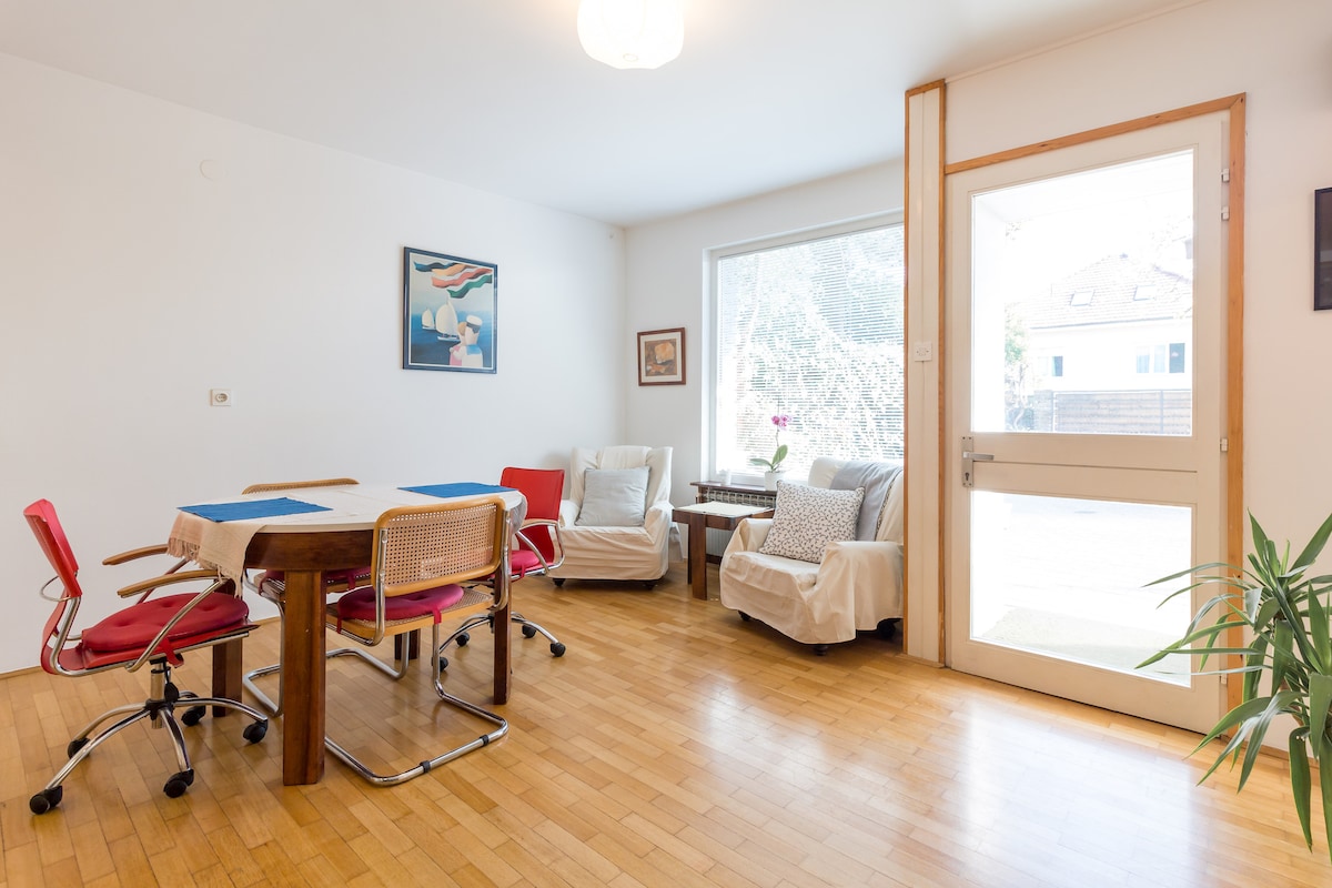A bright living space features a wooden table surrounded by four chairs, complemented by two cozy armchairs. A large window allows natural light to fill the room, and a small table with a potted plant is positioned beside the seating area, creating an inviting atmosphere.