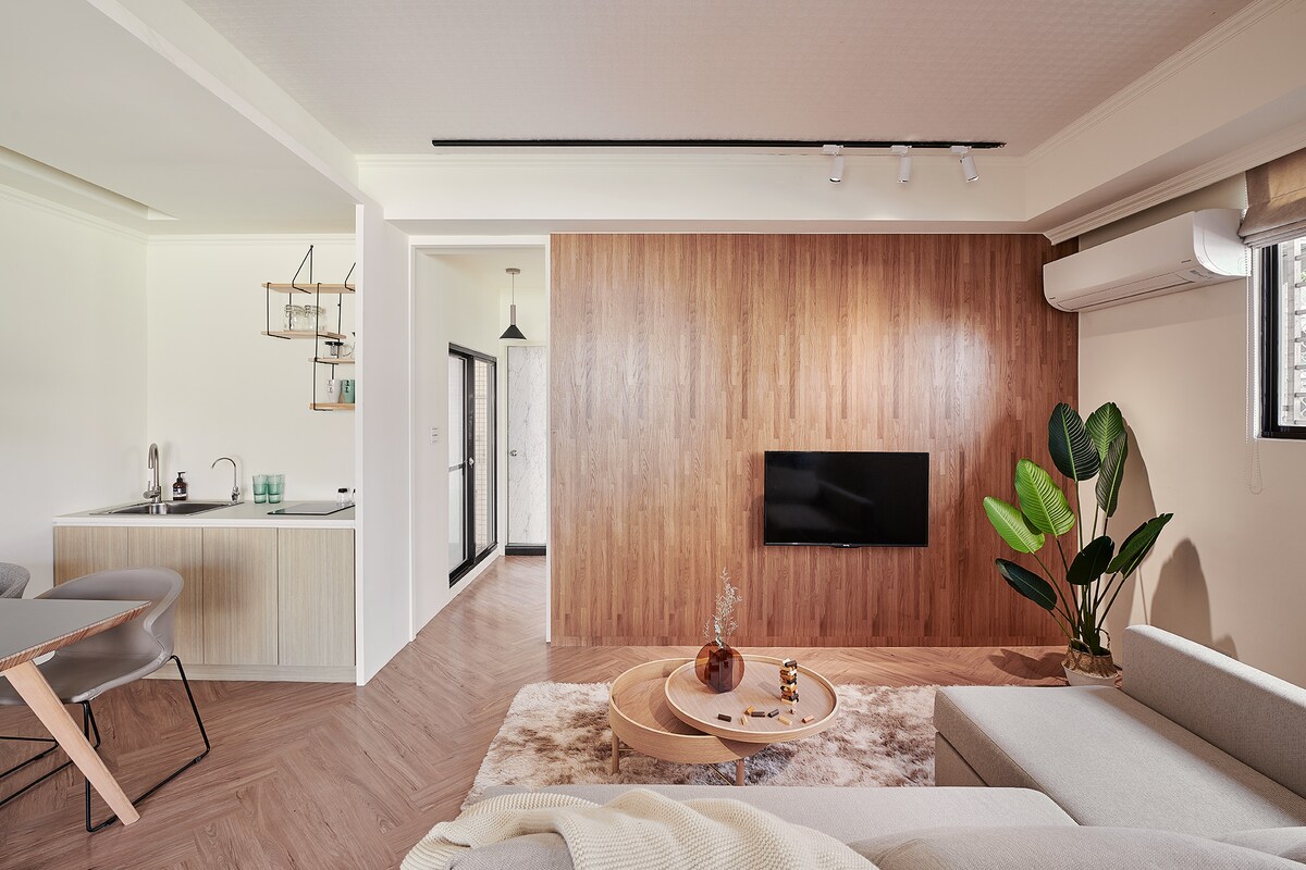 A modern living area features a light-colored sofa, a circular coffee table, and a mounted television against a wooden accent wall. A dining area with sleek chairs is visible, along with a well-equipped kitchen space in the background. Natural light enters through a window beside indoor plants.
