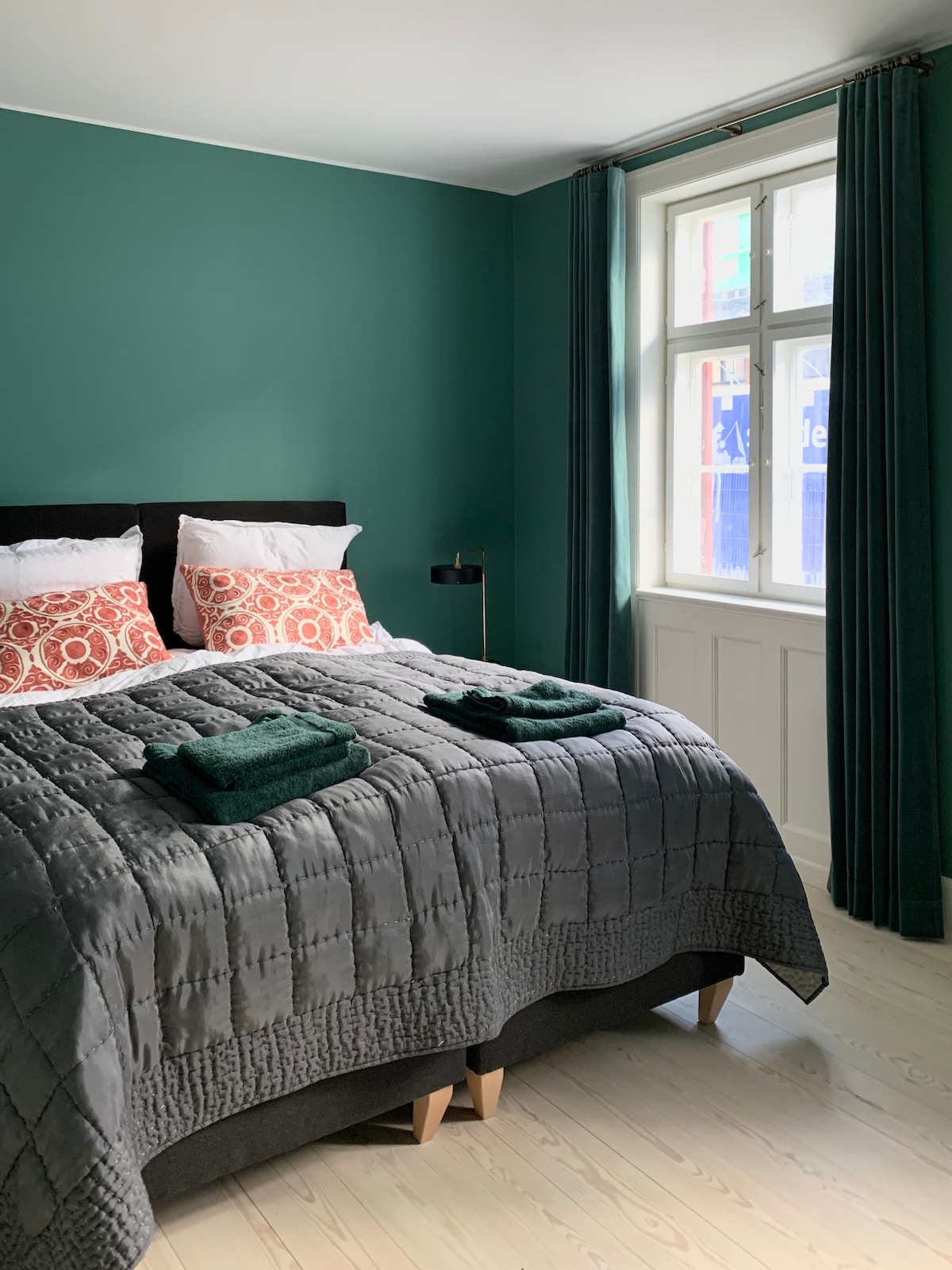 A spacious bedroom is presented with green walls and a large window allowing natural light. A dark quilted bedspread covers a generous bed, adorned with two patterned pillows. Two rolled towels in dark green lie on the bed, while matching curtains frame the window.