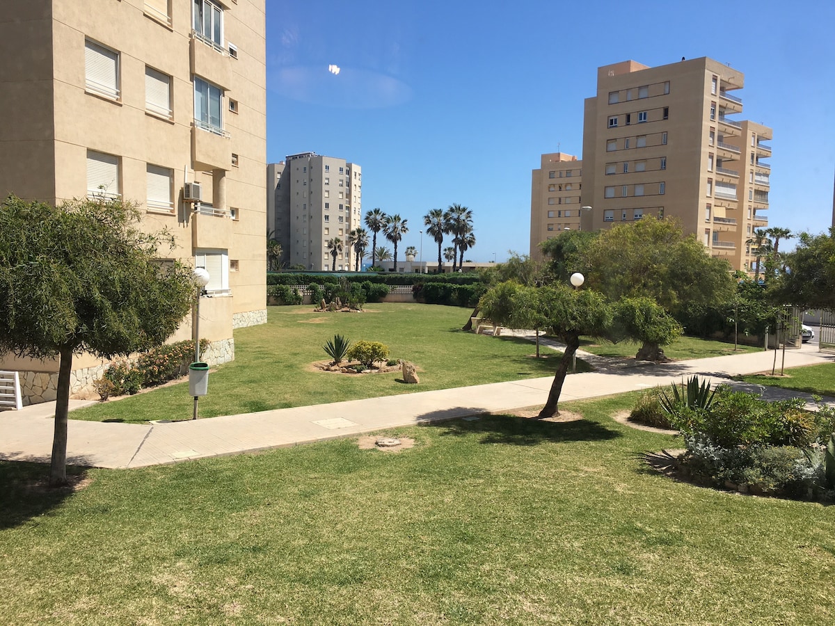 A green lawn is surrounded by landscaped gardens, featuring a variety of shrubs and trees. Buildings are visible in the background, with a clear blue sky above. Pathways are lined with light fixtures, providing a welcoming outdoor space near the apartment.