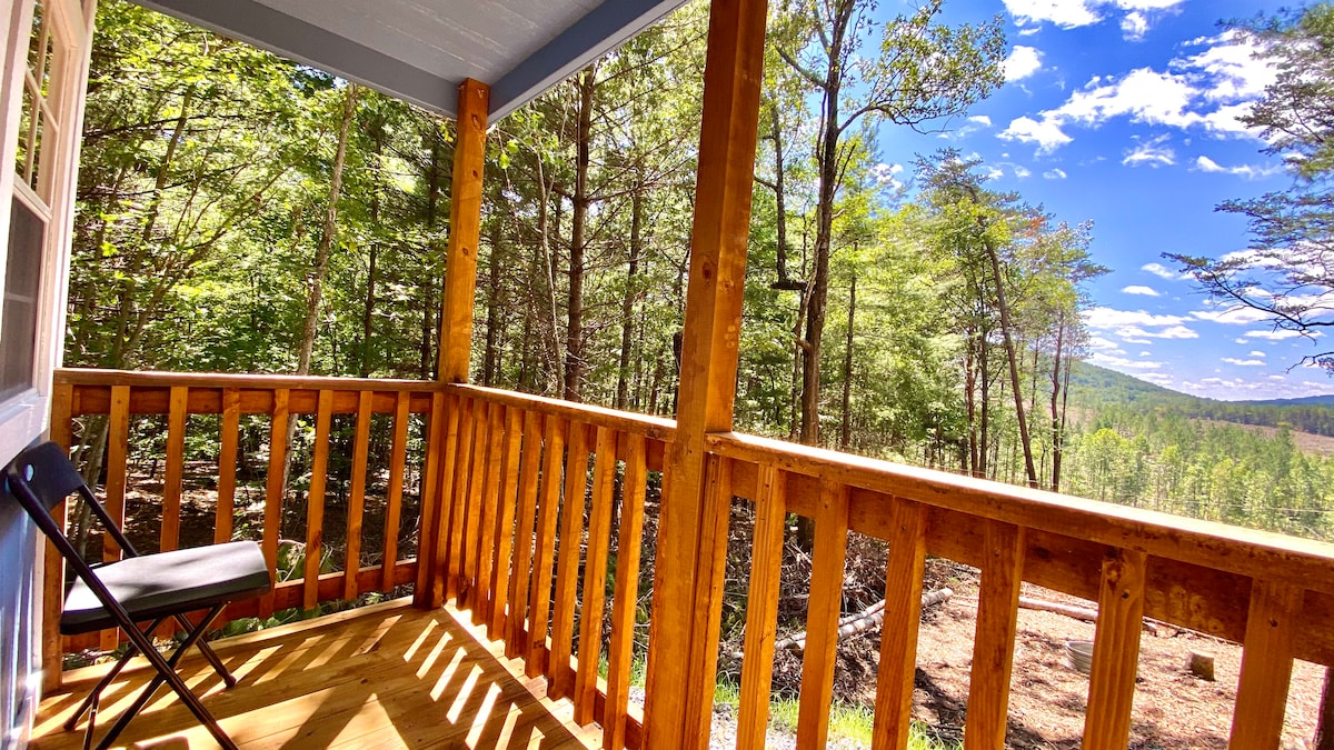 A wooden deck offers a serene view of the surrounding woods and distant mountains. Sunlight filters through the trees, illuminating the space. A black folding chair is positioned on the deck, inviting relaxation while enjoying nature.