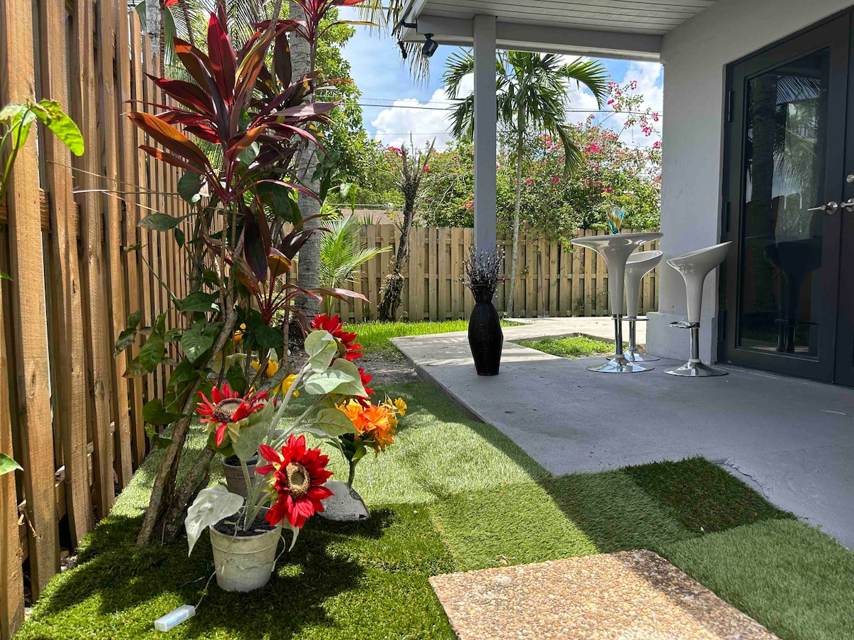 A landscaped outdoor area is presented, featuring vibrant flowers in planters and artificial grass. Two modern chairs accompany a small white table, while a slender vase adds an elegant touch. The space is surrounded by natural greenery and a wooden fence, providing a sense of privacy.