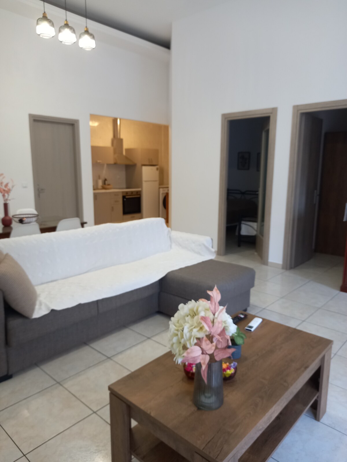 A spacious living area is shown, featuring a comfortable gray sofa adorned with a light throw. A wooden coffee table is in the center, decorated with flowers. The kitchen area is visible in the background, alongside an open doorway leading to another room.