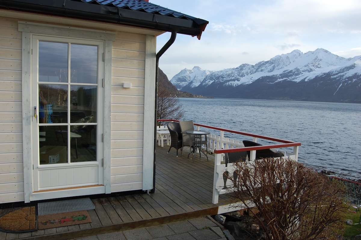 A wooden cabin exterior is shown, with a glass door leading to a spacious deck overlooking the water. Patio furniture is arranged on the deck, complemented by a stunning view of the mountains in the background, enhancing the cabin's serene location.