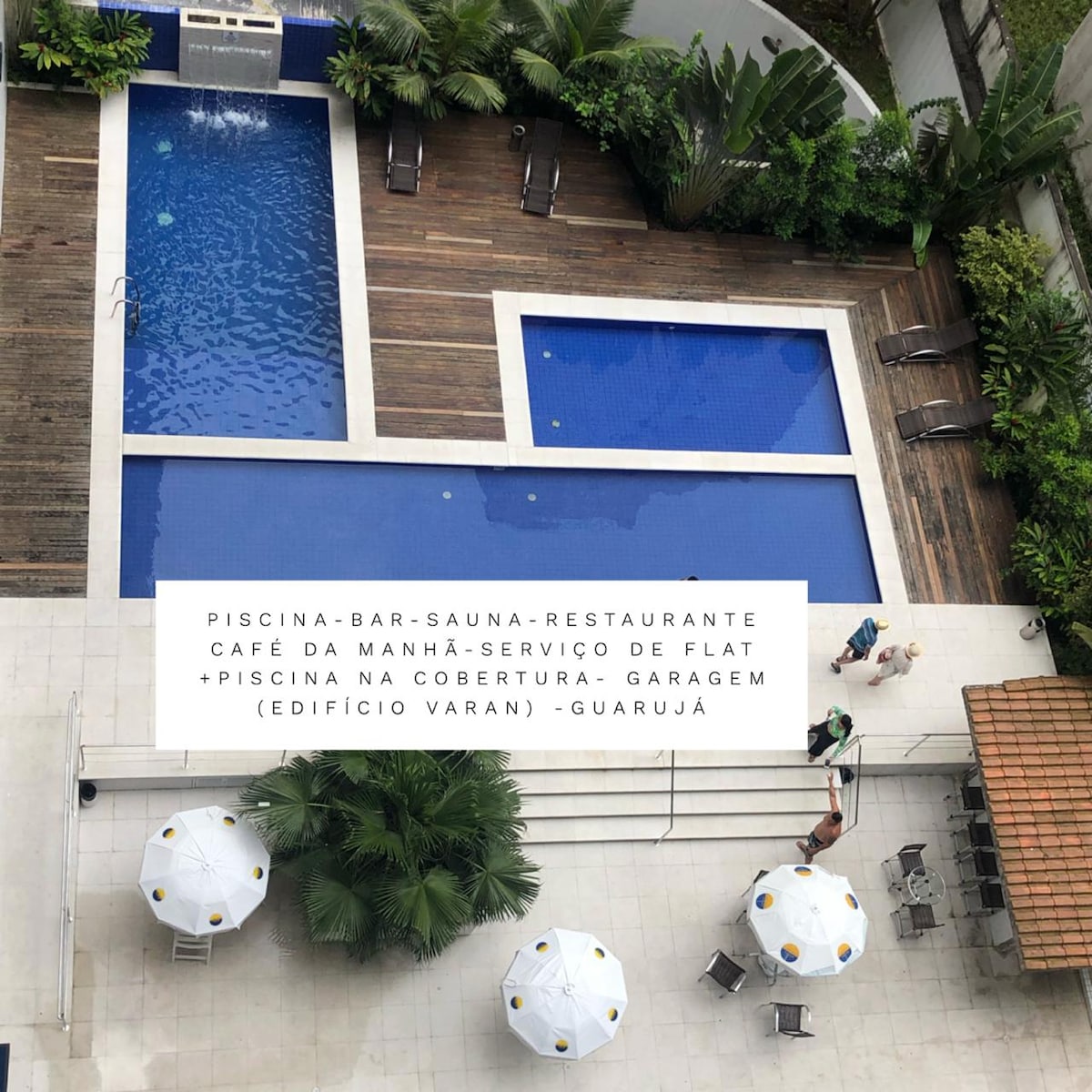 Two separate swimming pools are surrounded by wooden decking, with sun loungers arranged nearby. Lush greenery enhances the setting. A cafe and restaurant area can be seen adjacent to the pools, offering seating under white umbrellas for shade.