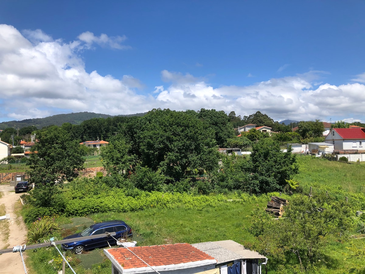 A panoramic view of the surrounding landscape showcases lush greenery, with a mix of trees, shrubs, and open fields. In the foreground, a gravel path leads toward a cluster of buildings. The sky is bright blue with scattered clouds, enhancing the natural scenery.