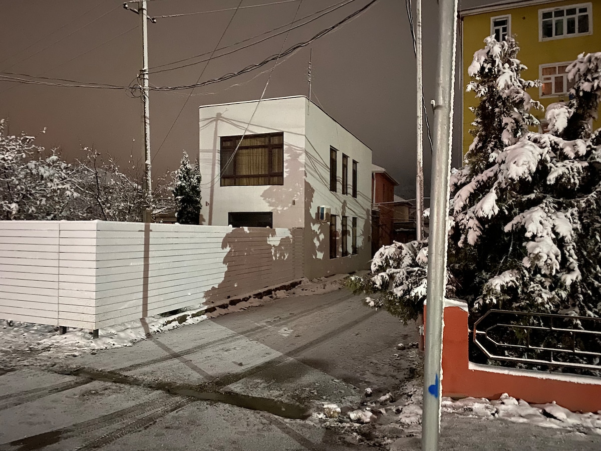 A modern, two-story home is captured under soft nighttime lighting, with snow covering the ground and surrounding areas. The exterior features large windows and a simple facade. Nearby trees and a white fence create a tranquil setting, while the street is partially visible coated in snow.