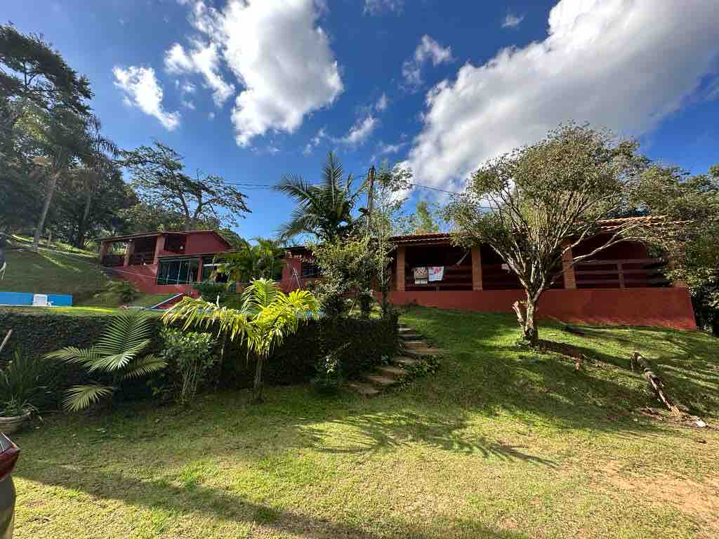 A spacious property is set against a backdrop of greenery and trees, featuring well-maintained gardens. The main structure displays an outdoor porch with a railing, inviting views of the surrounding landscape. A clear blue sky and soft clouds add a serene atmosphere to the setting.
