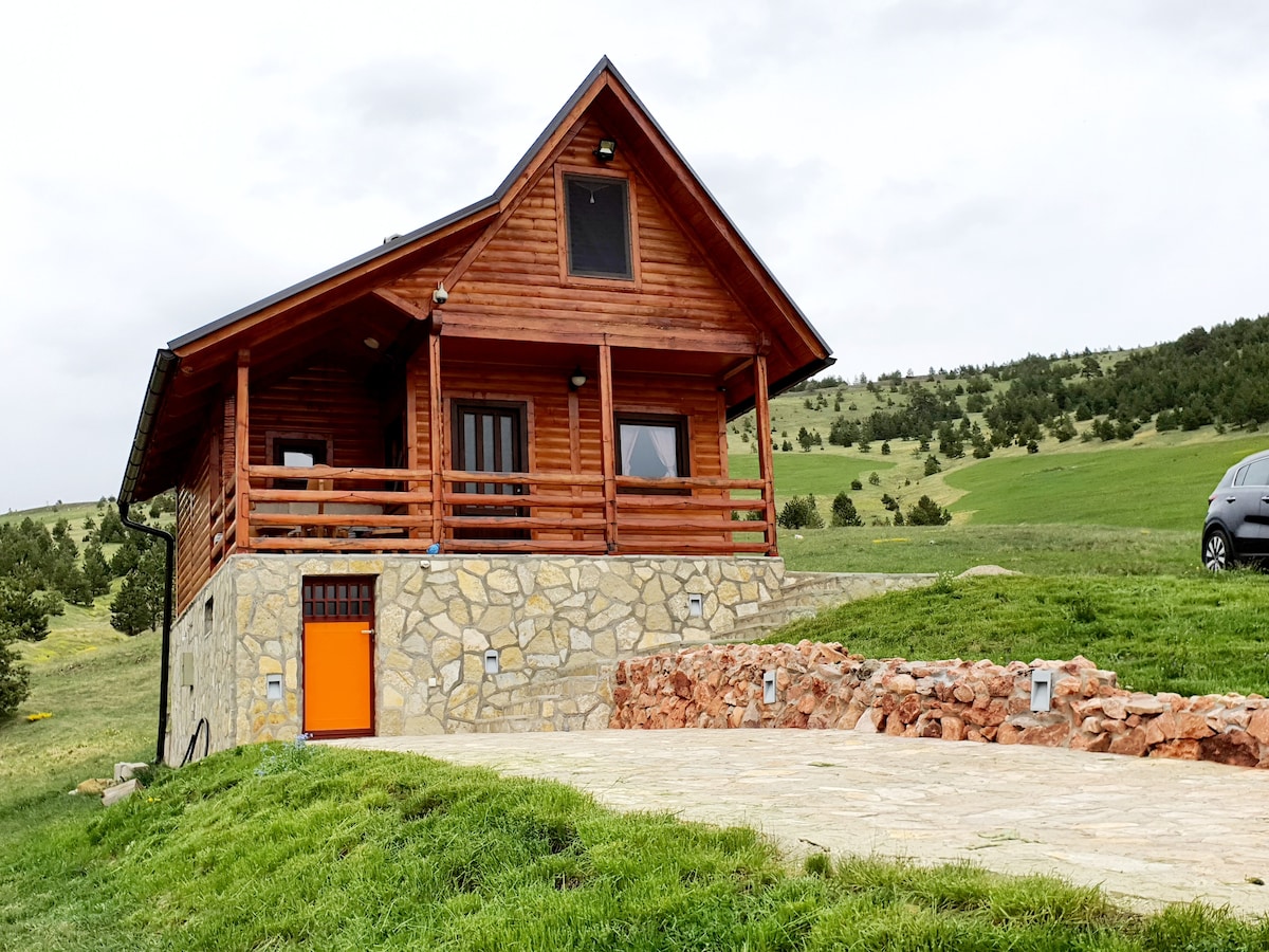 A wooden lodge featuring a sloped roof and a welcoming porch with wooden railings. The stone base adds texture to the structure, and an orange door provides a vibrant accent. Lush green hills are visible in the background, enhancing the serene outdoor setting.