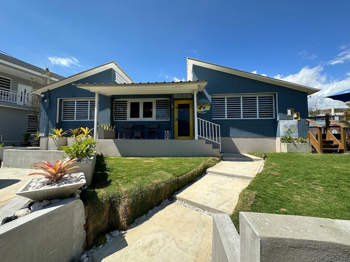 A charming blue house is set against a clear blue sky, featuring a welcoming yellow door. The well-kept front yard displays a mix of greenery and decorative plants, while a walkway leads to the entrance. The two gabled rooflines create an inviting appearance.