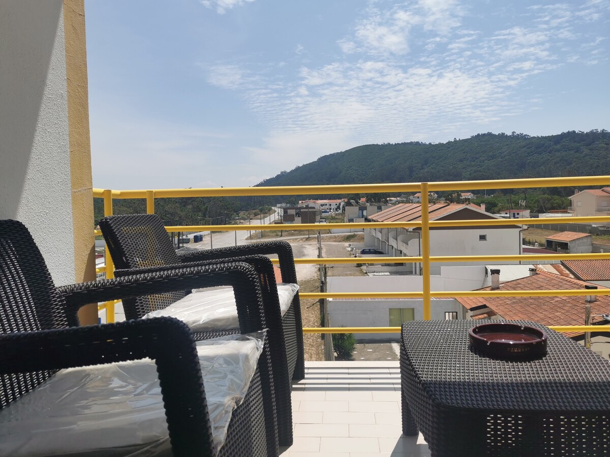 A balcony features outdoor furniture, including two chairs and a small table. The view includes a distant mountain and surrounding buildings. A warm sunlight casts soft shadows, enhancing the open space while providing an inviting spot for relaxation.