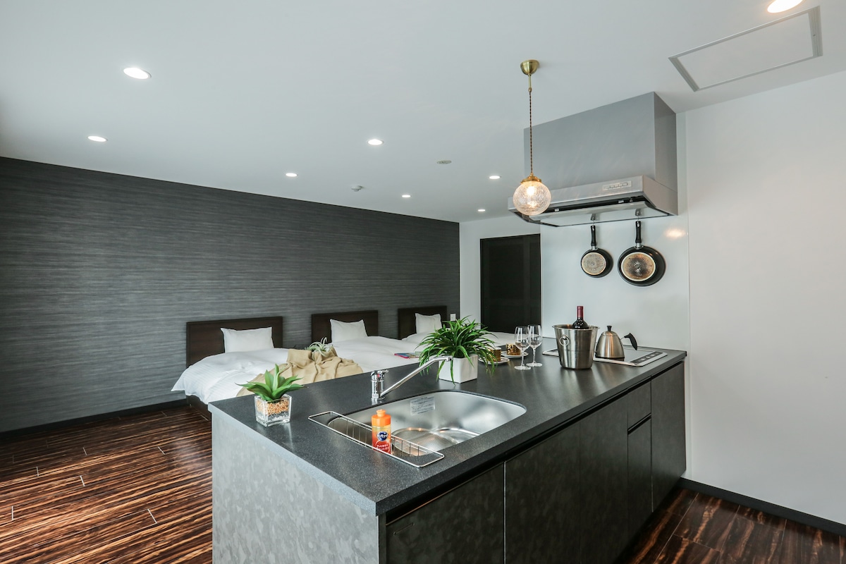 The room features a modern kitchen island with a sleek sink and minimalistic decor. A warm-toned living area is highlighted by two spacious beds in the background. Stylish pendant lighting hangs overhead, enhancing the contemporary design.