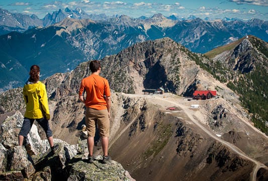 Two individuals are seen standing on rocky terrain, gazing at the expansive mountain landscape. The foreground features jagged rocks, while the backdrop showcases a range of mountains under a clear blue sky, with a small building visible in the distance.