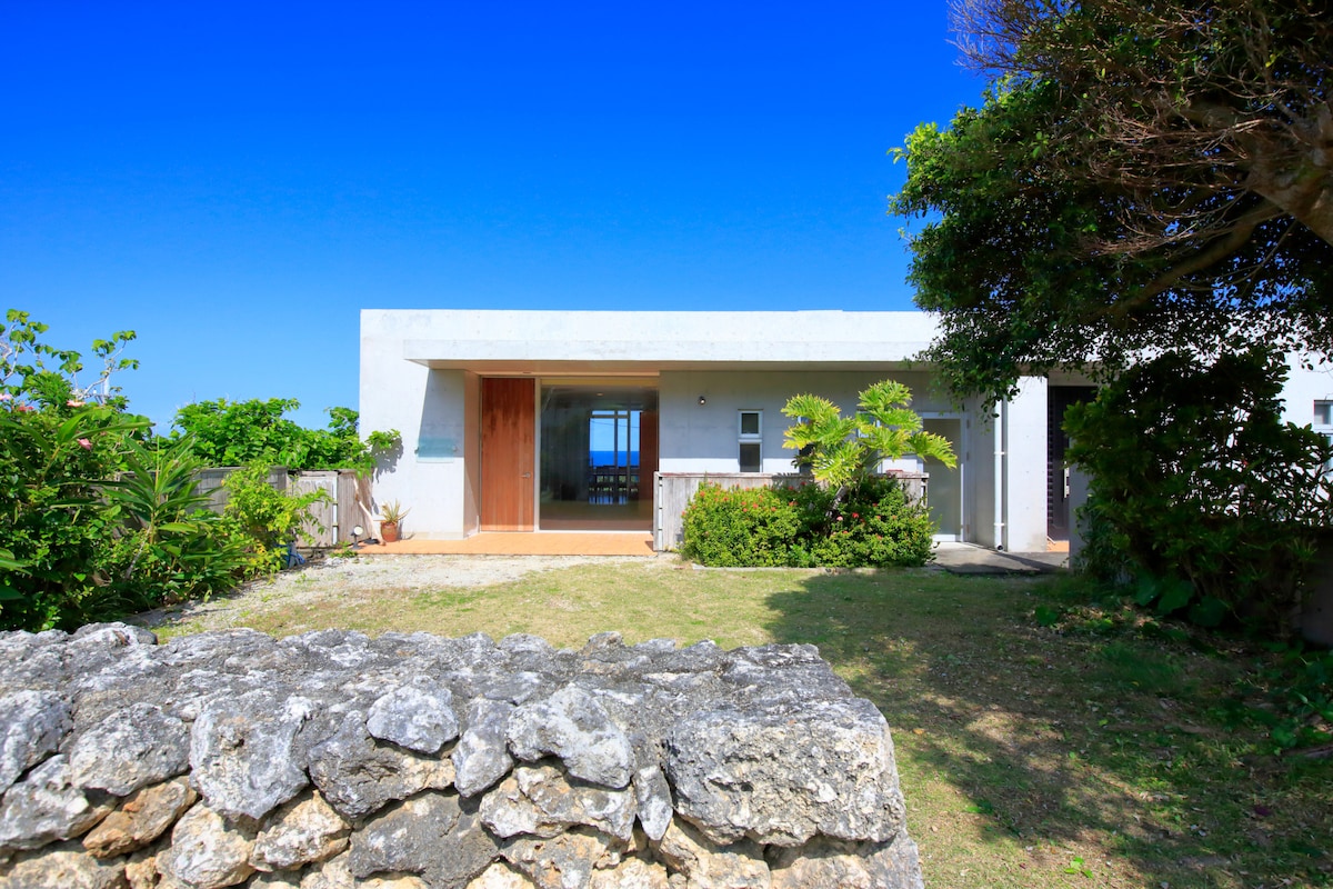 The exterior of the property features a modern structure set against a clear blue sky. Lush greenery surrounds the entrance, complemented by a subtle stone wall. The doorway leads to an inviting space, seamlessly connecting indoor and outdoor areas.