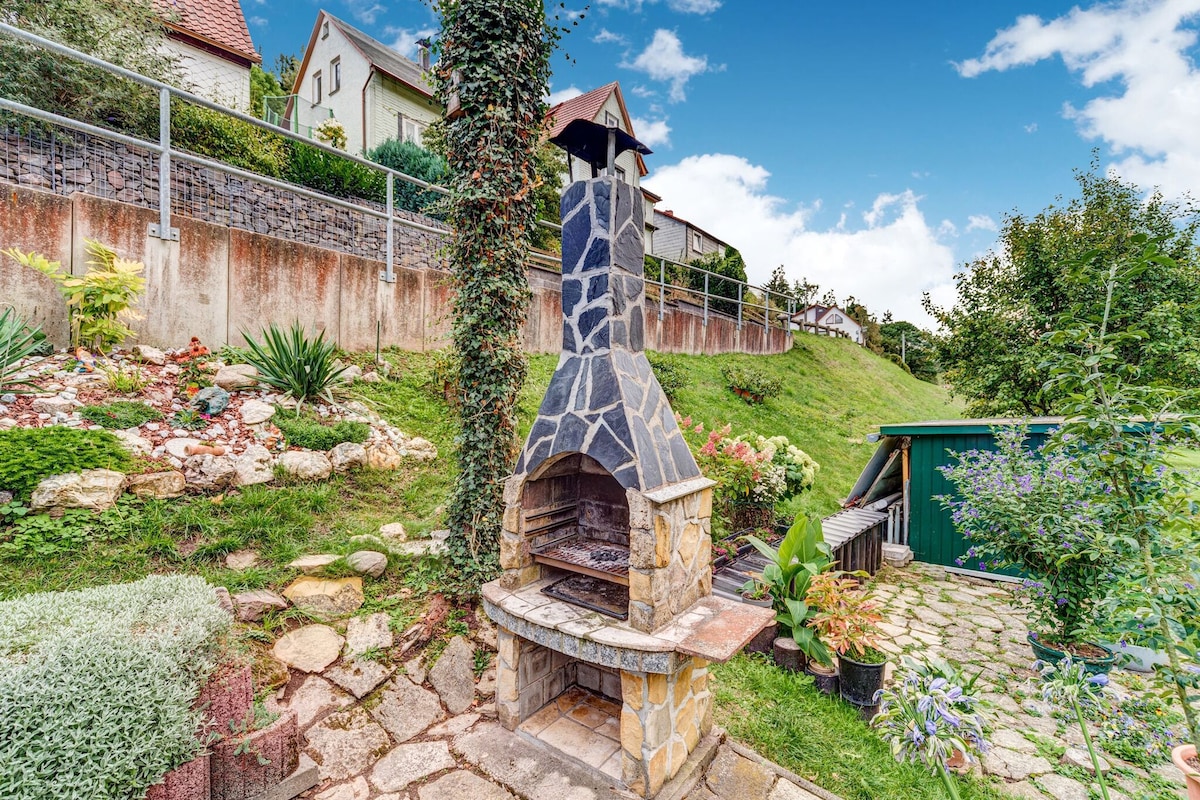 A stone barbecue area is situated amidst a lush garden, featuring a stacked stone design and a chimney. Surrounding greenery and colorful plants enhance the outdoor setting, while a paved walkway leads up to the grill, inviting outdoor cooking and relaxation.