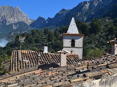 The image showcases the rooftops of a rustic village, highlighted by traditional tiled roofs. A church tower rises prominently against a backdrop of rugged mountains under a clear blue sky. The natural landscape and architectural elements blend harmoniously in the serene environment.