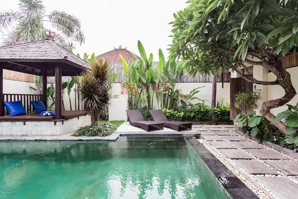 A serene pool area is surrounded by lush tropical greenery. Two lounge chairs are positioned at the water's edge, while a gazebo with blue cushions offers relaxation. The pathway is lined with stone tiles and decorative plants, creating a tranquil outdoor space.