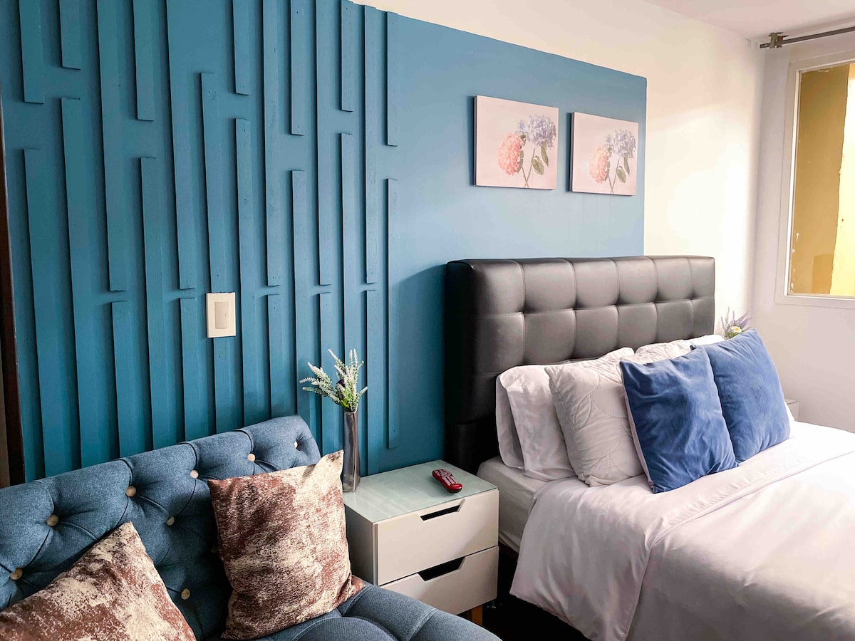 A modern sleeping area features a stylish blue accent wall adorned with floral artwork. A spacious bed with plush pillows is complemented by a cozy seating area with decorative cushions. Natural light enters through a nearby window, enhancing the inviting ambiance.