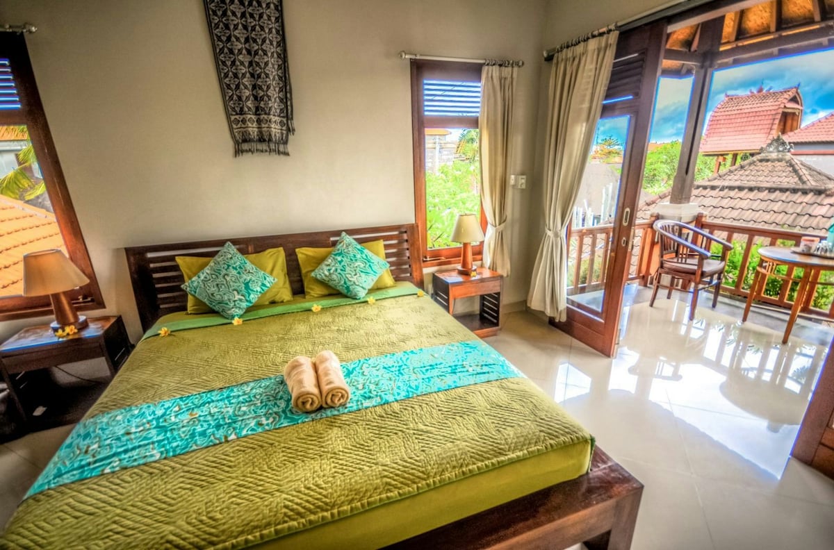 A spacious room features a queen-sized bed adorned with decorative pillows. Natural light filters through large windows, casting reflections on the glossy floor. A small dining area is visible in the background, complemented by a touch of greenery from the surroundings.