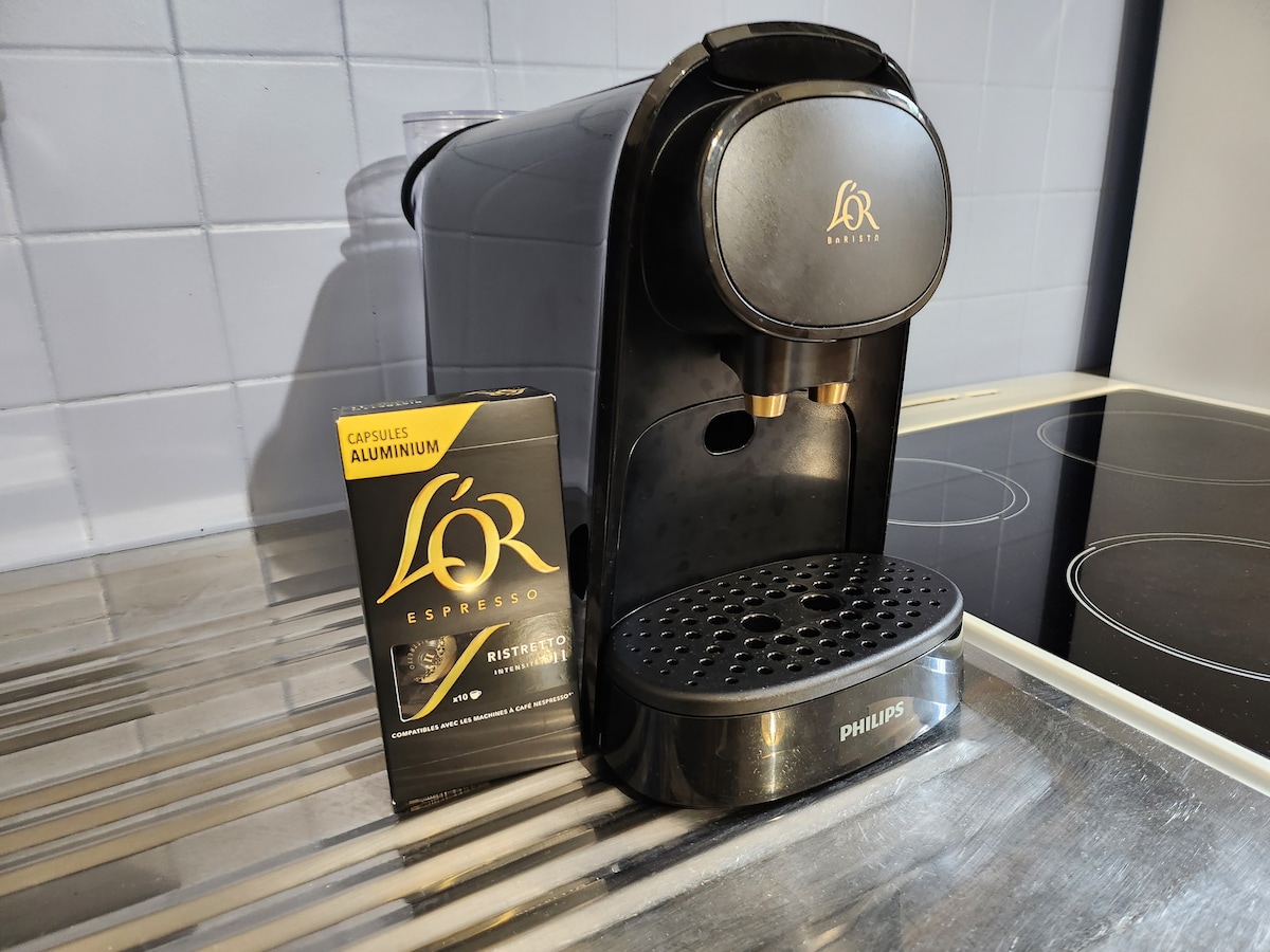 A sleek black espresso machine is positioned on a countertop, accompanied by a box of aluminum coffee capsules. The machine features two spouts for brewing, with a drip tray below for convenience. The backsplash displays a textured pattern, adding a modern touch.