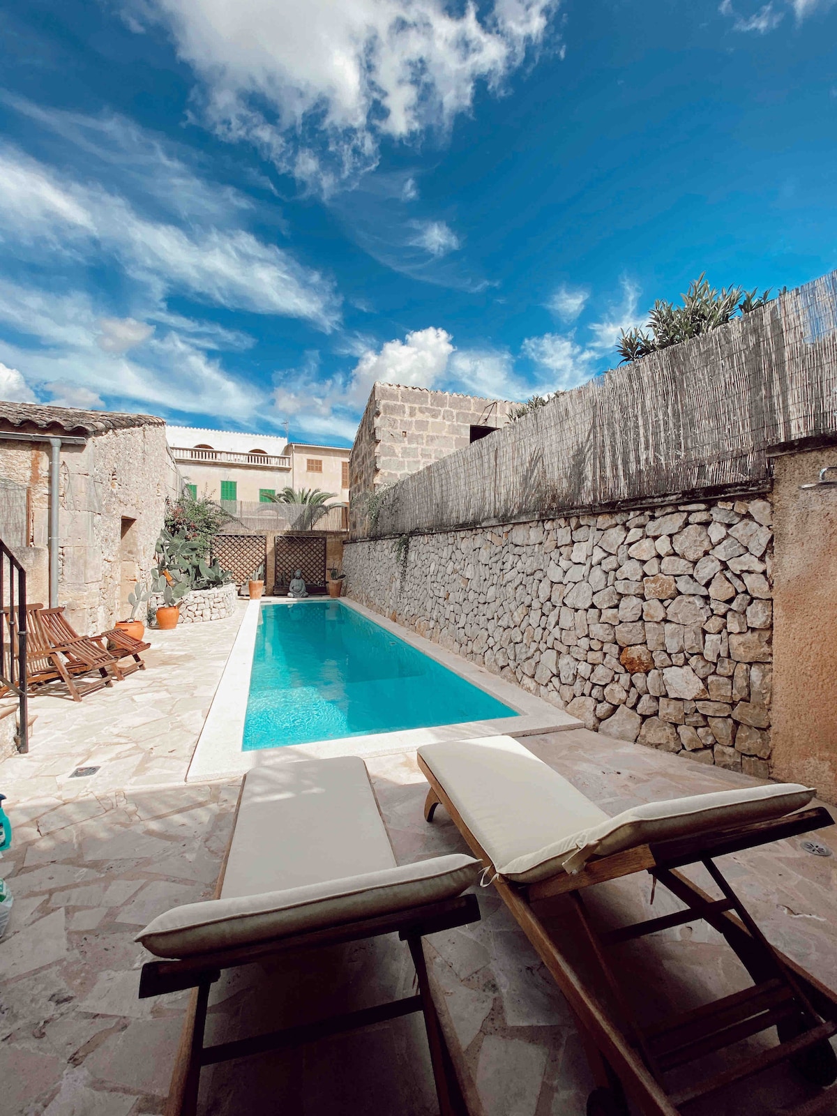 A serene outdoor area features a clear swimming pool bordered by natural stone walls. Two lounge chairs are positioned nearby on a stone patio, with greenery visible in the background, and a bright blue sky overhead enhances the tranquil setting.