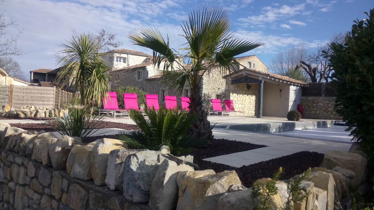 A landscaped outdoor area features a private swimming pool and lounge chairs lined in vibrant pink. Surrounding greenery includes palm trees and decorative stones, adding a touch of color and texture to the space. The stone wall provides a rustic backdrop to this tranquil environment.