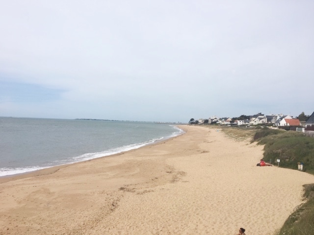 A sandy beach stretches along the coastline, meeting the gentle waves of the sea. Residential buildings are visible in the distance along the shore. The sky is lightly overcast, creating a calm and serene atmosphere.