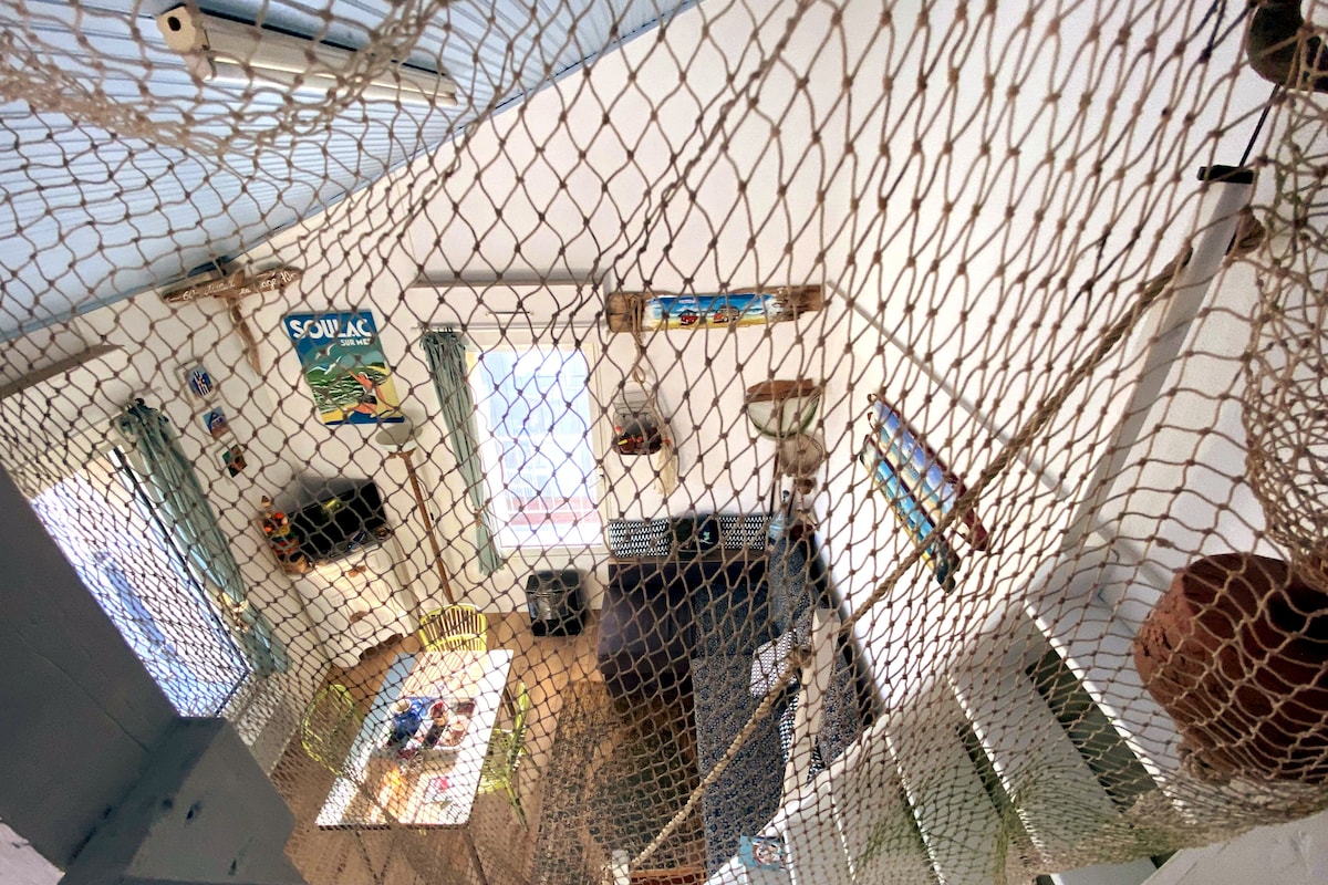 An aerial view of the living area features a fishing net overhead, providing a unique texture. The bright, open space includes a dining table surrounded by colorful chairs and a cozy seating area. Light floods through large windows, enhancing the airy ambiance.