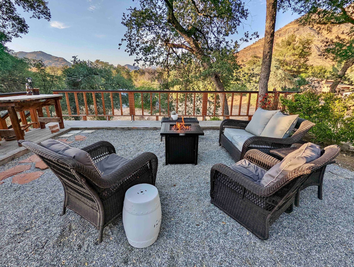 A cozy outdoor seating area is arranged with two wicker chairs, a smooth table, and a central fire pit. Natural surroundings are highlighted by the oak trees and scenic mountain views, creating a welcoming space for relaxation and socializing.