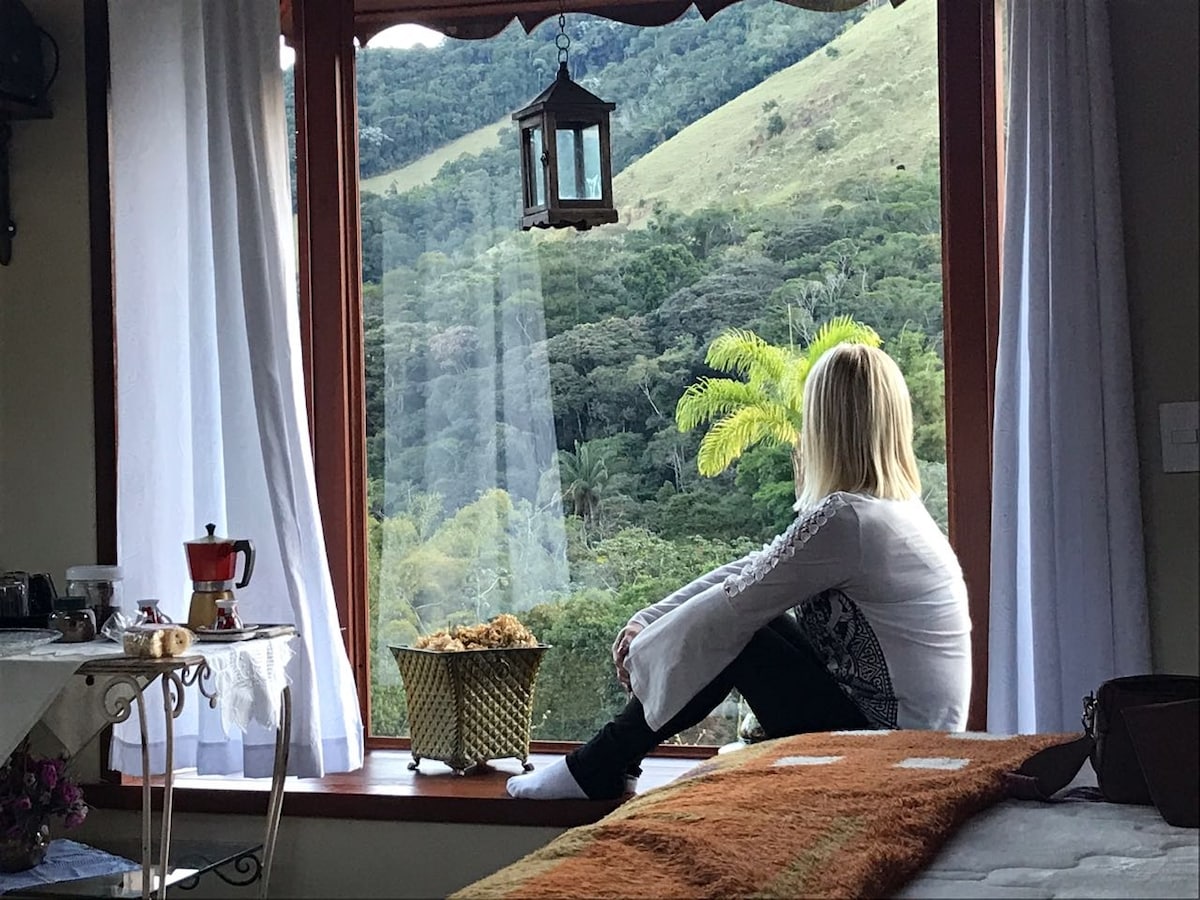 A bay window offers a scenic view of lush greenery and rolling hills. Soft white curtains frame the window, and a small table sits nearby with a coffee pot and treats. A cozy blanket rests on the bed, inviting relaxation in this serene setting.