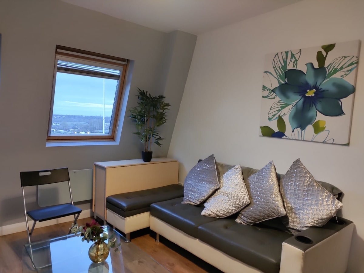 A modern living area features a comfortable sofa adorned with decorative pillows. A sleek coffee table rests on polished wooden flooring. A minimalist chair is positioned nearby, while a vibrant wall art piece and a potted plant bring a touch of nature to the space.