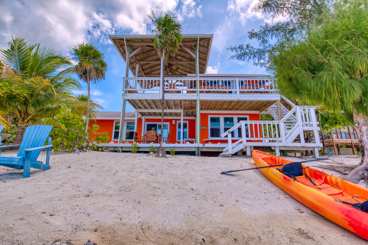 A charming oceanfront house features a wooden deck with an overhang and ample seating. The vibrant exterior is complemented by lush greenery and sandy terrain. An orange kayak rests on the beach, while beach chairs invite relaxation under a partly cloudy sky.