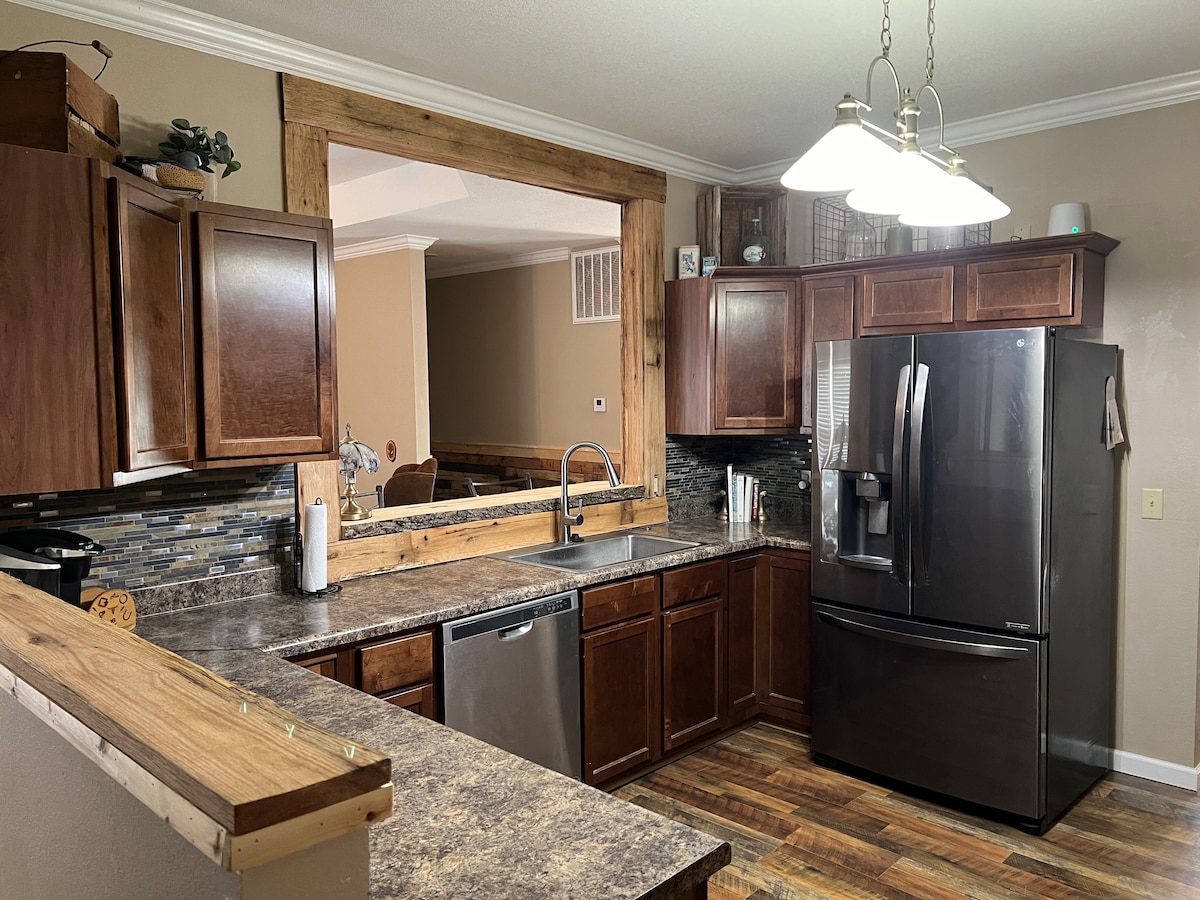 The kitchen features wooden cabinets and a spacious countertop with a contemporary design. A modern refrigerator is positioned beside a stainless steel sink, and three pendant lights provide illumination. The open layout allows for easy access to the adjoining living space.