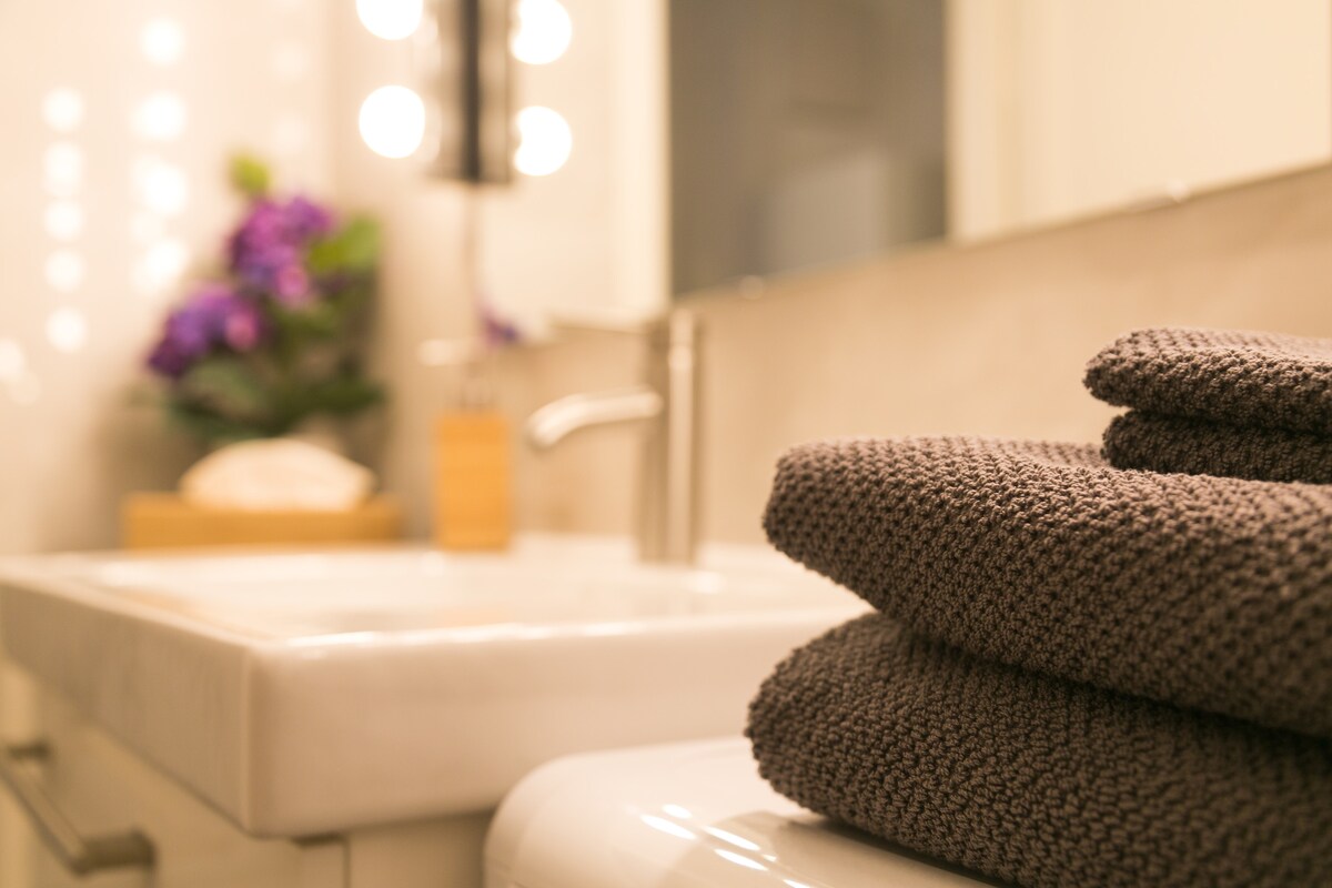 A modern bathroom showcases neatly stacked dark towels positioned beside a sleek, white sink. Soft lighting from an illuminated mirror highlights the clean lines and neutral palette. A touch of greenery can be seen in the background, adding a subtle decorative element.