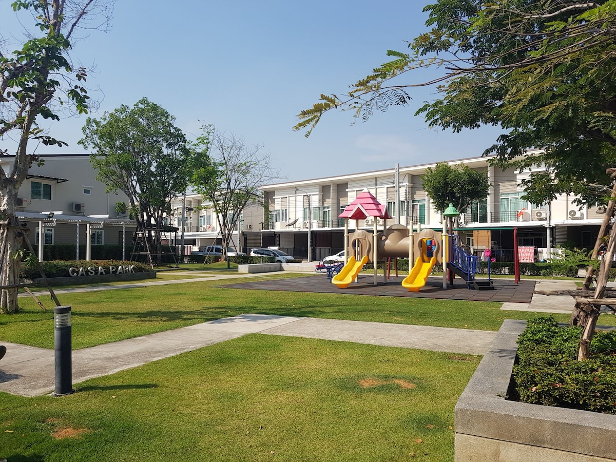 A spacious playground area is featured with colorful play structures, including slides and swings. Well-maintained grassy areas surround the playground, complemented by trees providing shade. Nearby, a path leads to residential buildings, creating a tranquil and family-friendly atmosphere.