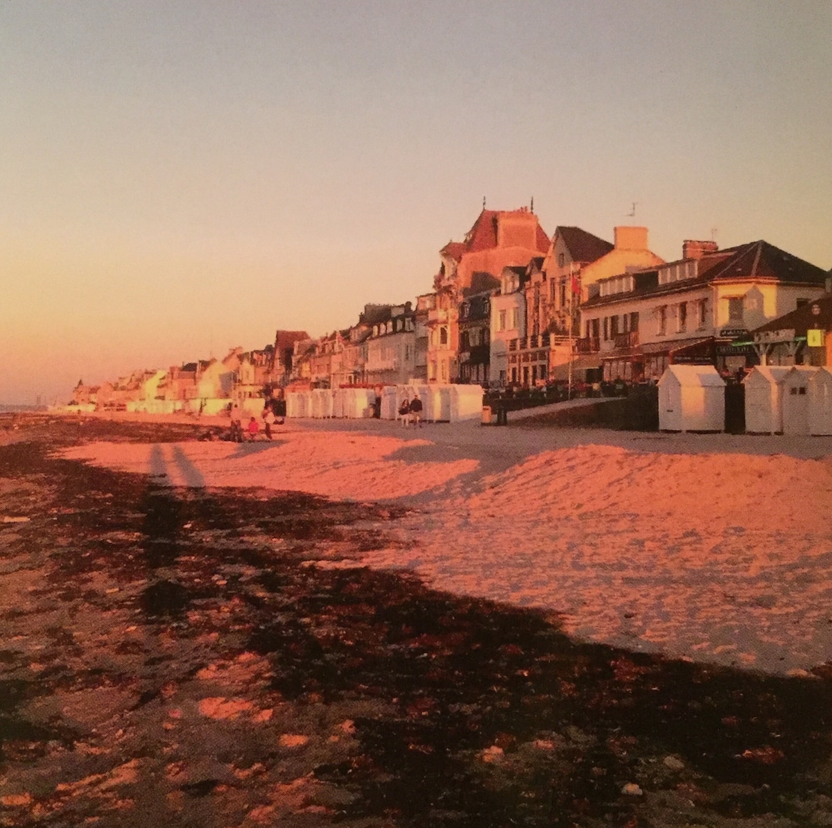 A sandy beach scene captured at sunset, featuring beach cabins lined along the shore. Shadows stretch across the sand, while buildings with architectural details provide a backdrop, indicating the presence of shops and cafes. The warm hues reflect off the sand, creating a tranquil atmosphere.