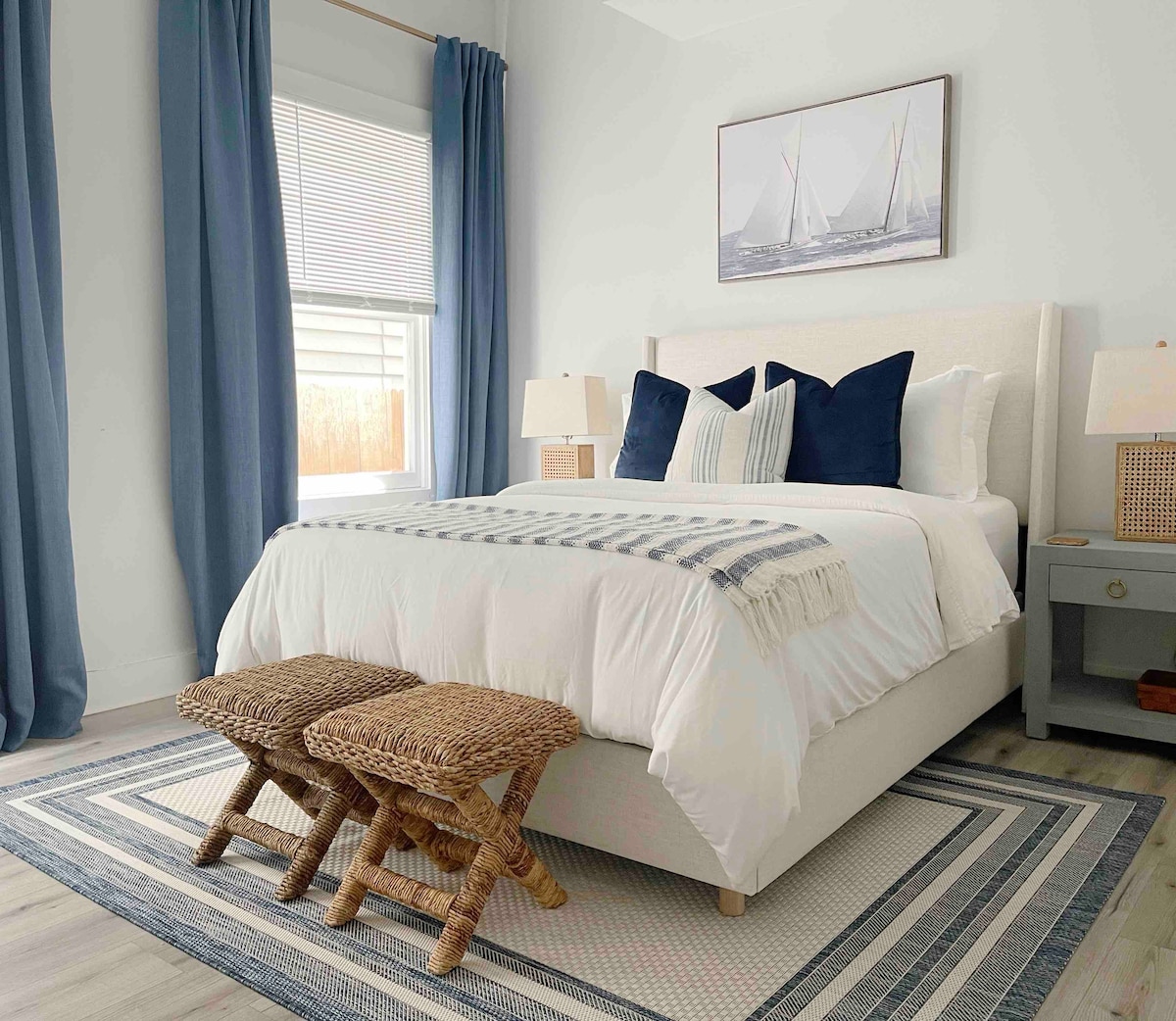 A serene bedroom features a spacious bed with white bedding and decorative pillows in shades of navy and cream. Two woven stools are placed at the foot of the bed, complementing the light-colored hardwood floor and area rug. Natural light enters through large windows, enhanced by blue curtain panels.