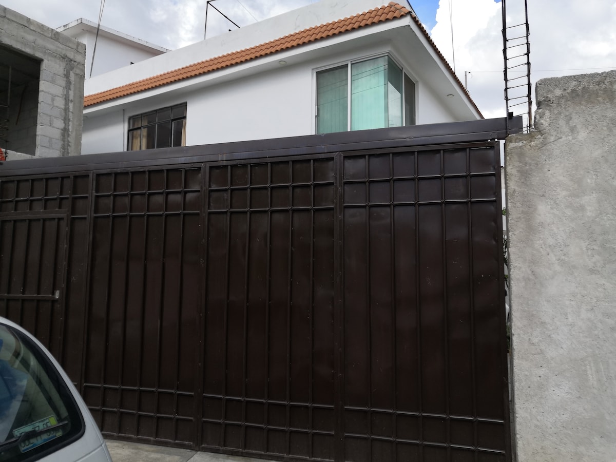 A solid brown metal gate provides entrance to the property, flanked by a grey concrete wall. Above, a white building with large windows is visible, featuring a sloped roof. The overall structure and surrounding area can be seen under a partly cloudy sky.
