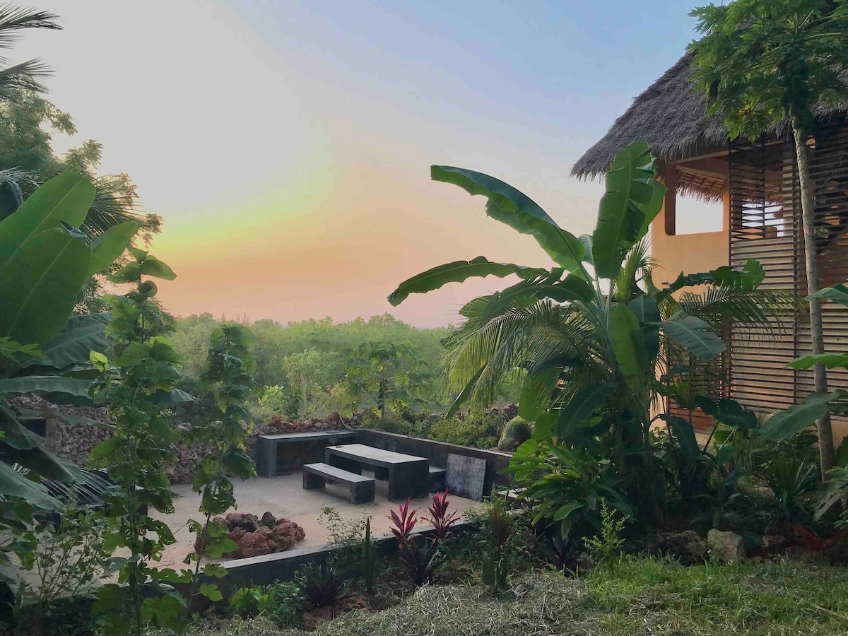 A tranquil outdoor seating area is surrounded by lush tropical plants, showcasing stone benches and a view of the vibrant landscape. The distant horizon reflects soft colors of the sunset, adding a serene ambiance to the space.