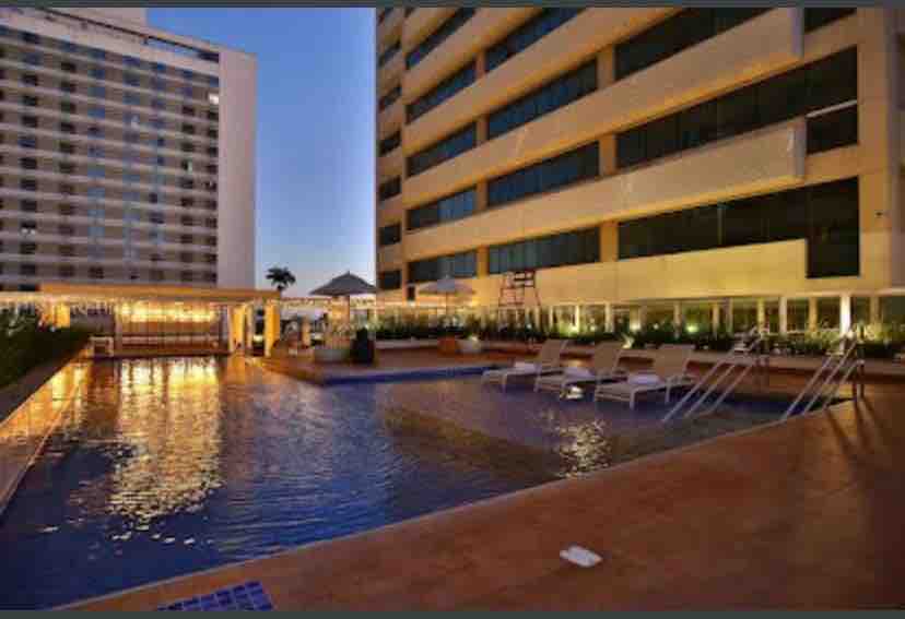 An outdoor pool reflects soft lighting, framed by modern buildings. Lounge chairs are positioned around the pool, providing space for relaxation. Lush greenery lines the area, contributing to a serene environment. The sunset casts a warm glow over the water.