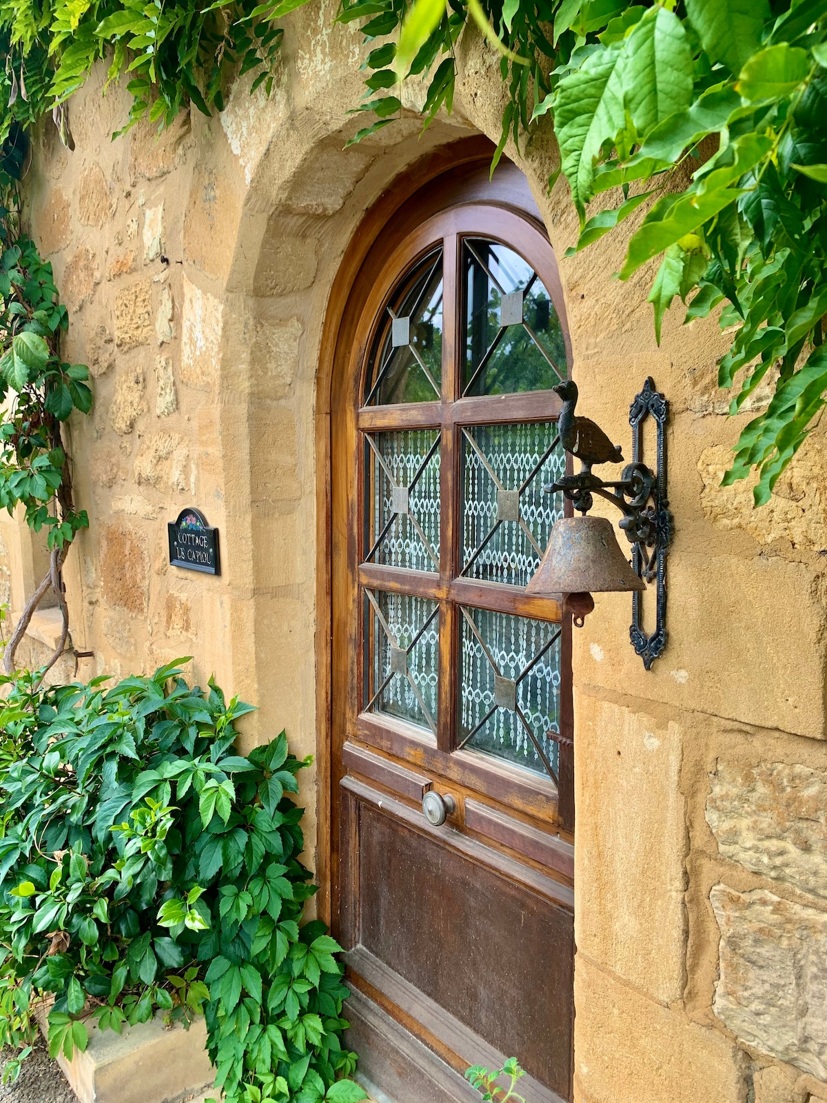 The entrance features a wooden door with decorative glass panels, framed by lush greenery and vibrant foliage. A vintage-style bell hangs beside the door, adding charm to the stone wall that surrounds the entrance.