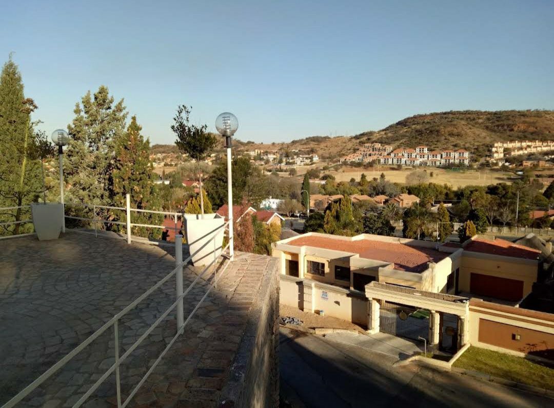 A spacious terrace area is shown, featuring a stone pathway and railing, overlooking a scenic view of nearby hills and residential structures. Soft lighting fixtures are visible, providing illumination for evening use.