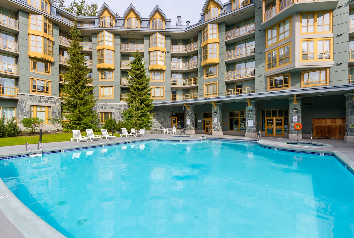 An outdoor pool is positioned in front of a four-story building with light gray siding and yellow accents. Surrounding the pool, several white lounge chairs are placed on a concrete deck, with tall evergreen trees providing a natural backdrop.