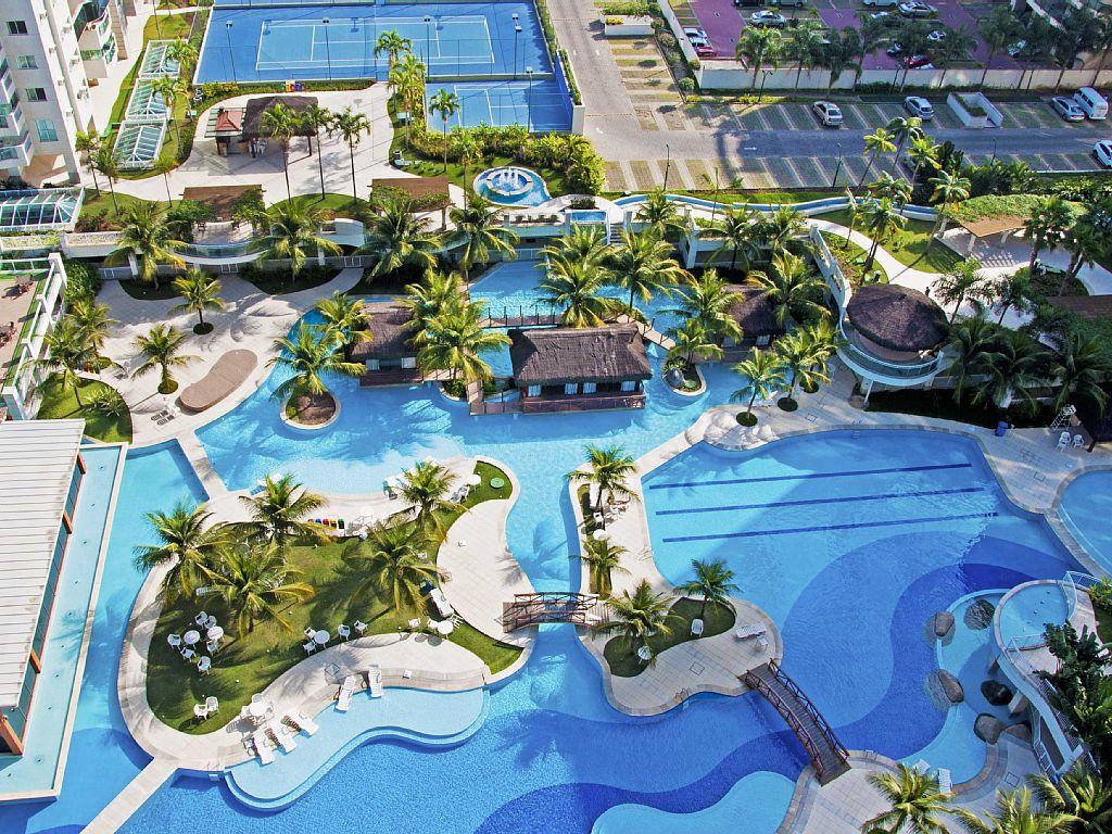 An expansive view showcases a resort-style complex featuring multiple swimming pools surrounded by palm trees. A central deck area includes a small building and various pathways connecting to landscaped gardens. Tennis courts are visible in the background, contributing to the recreational atmosphere.