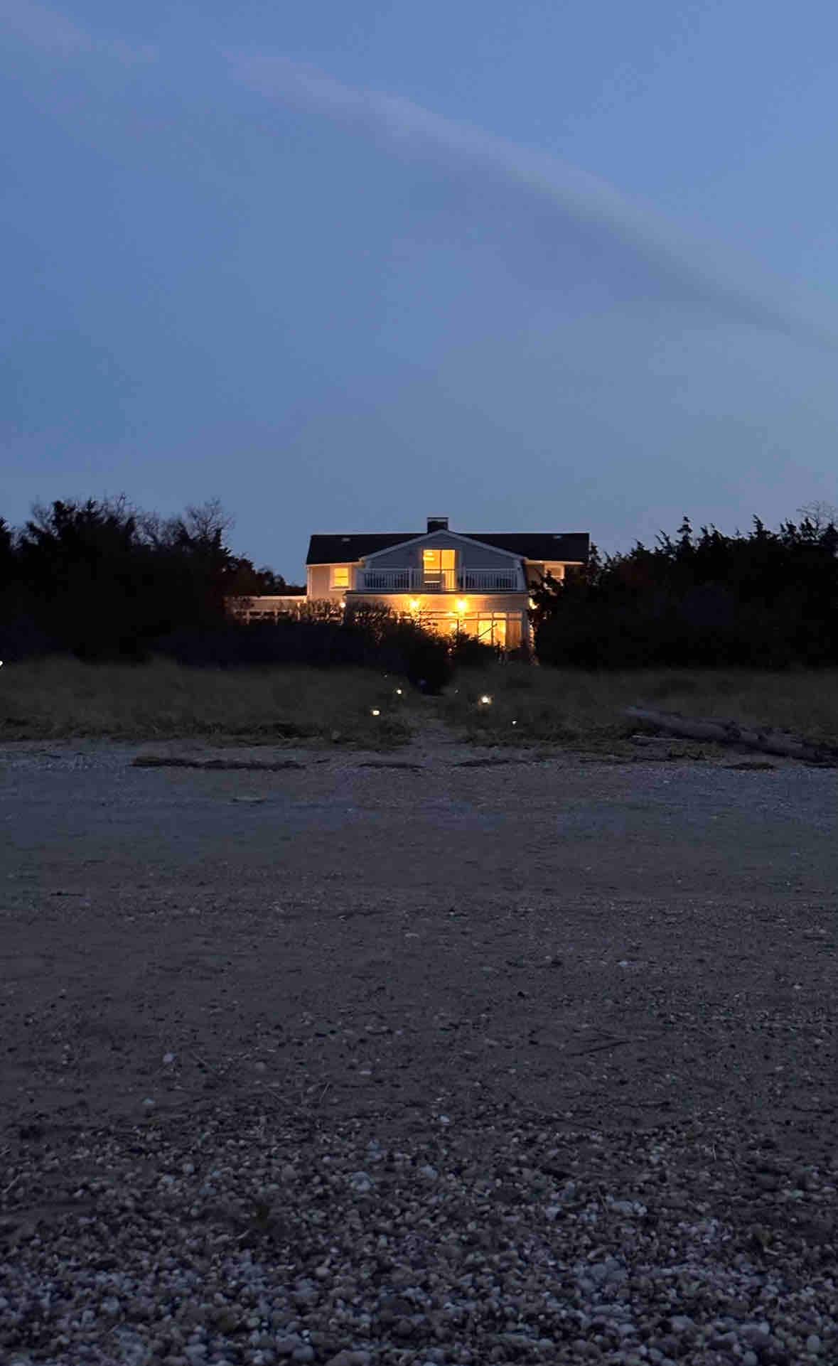 The exterior of a beachfront home is viewed at twilight, featuring warm illumination from windows that create a welcoming sight. Soft sand leads toward the house, framed by low vegetation. A sense of tranquility is conveyed by the calm evening ambiance.