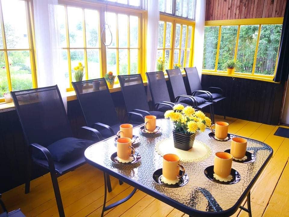 A bright sunroom features a large table set for breakfast, surrounded by several black chairs. A central vase holds yellow flowers, complemented by orange mugs and black plates, while ample natural light filters through large yellow-framed windows.