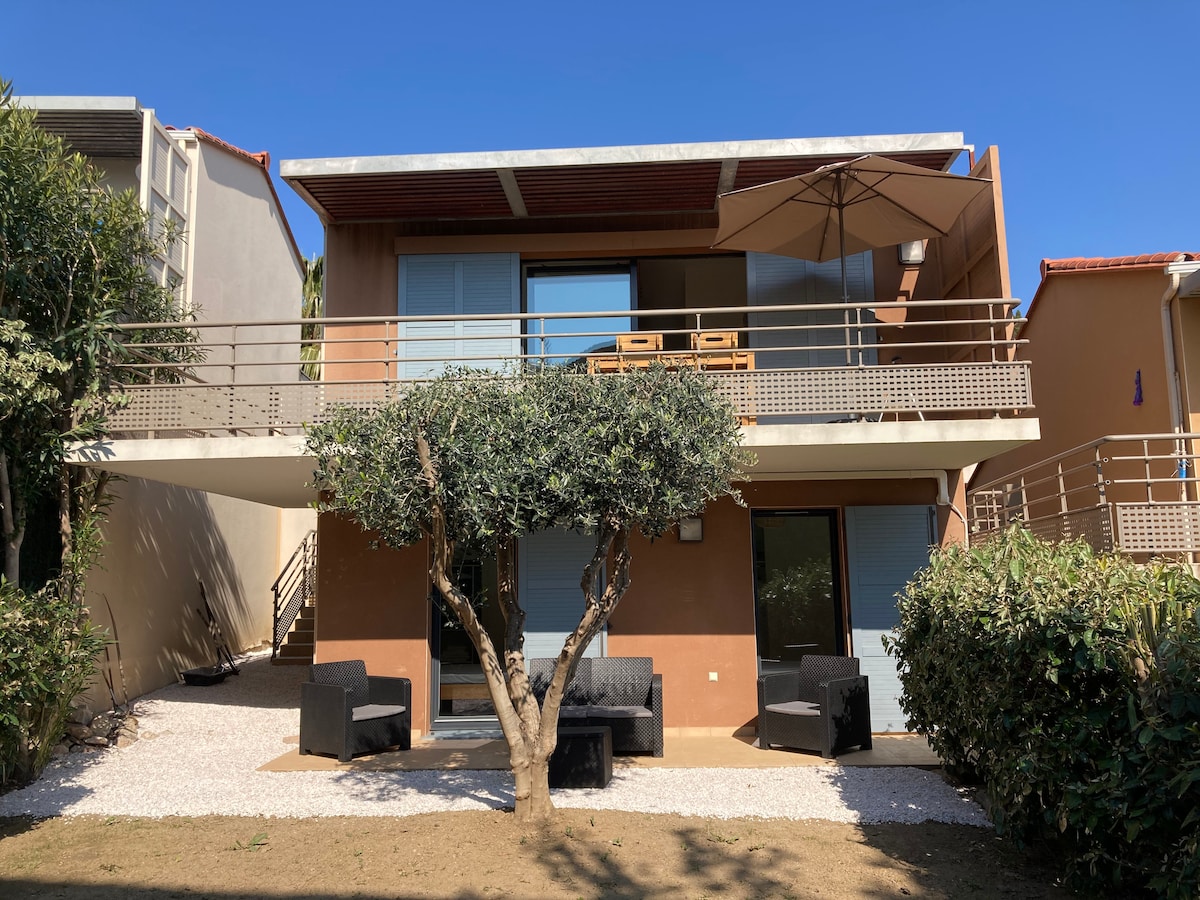 A modern villa is presented, featuring a spacious balcony with comfortable seating and a large umbrella. Lush greenery surrounds the property, with a tree positioned in the foreground. The architecture reflects contemporary style, complemented by clean lines and ample natural light.