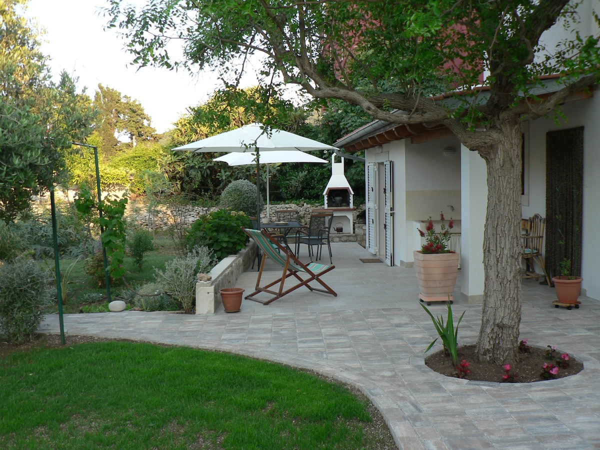 An outdoor space features a shaded seating area under a large umbrella, surrounded by landscaped greenery and flower pots. A barbecue grill stands in the corner, with stone pathways leading through the garden. A large tree provides additional shade.