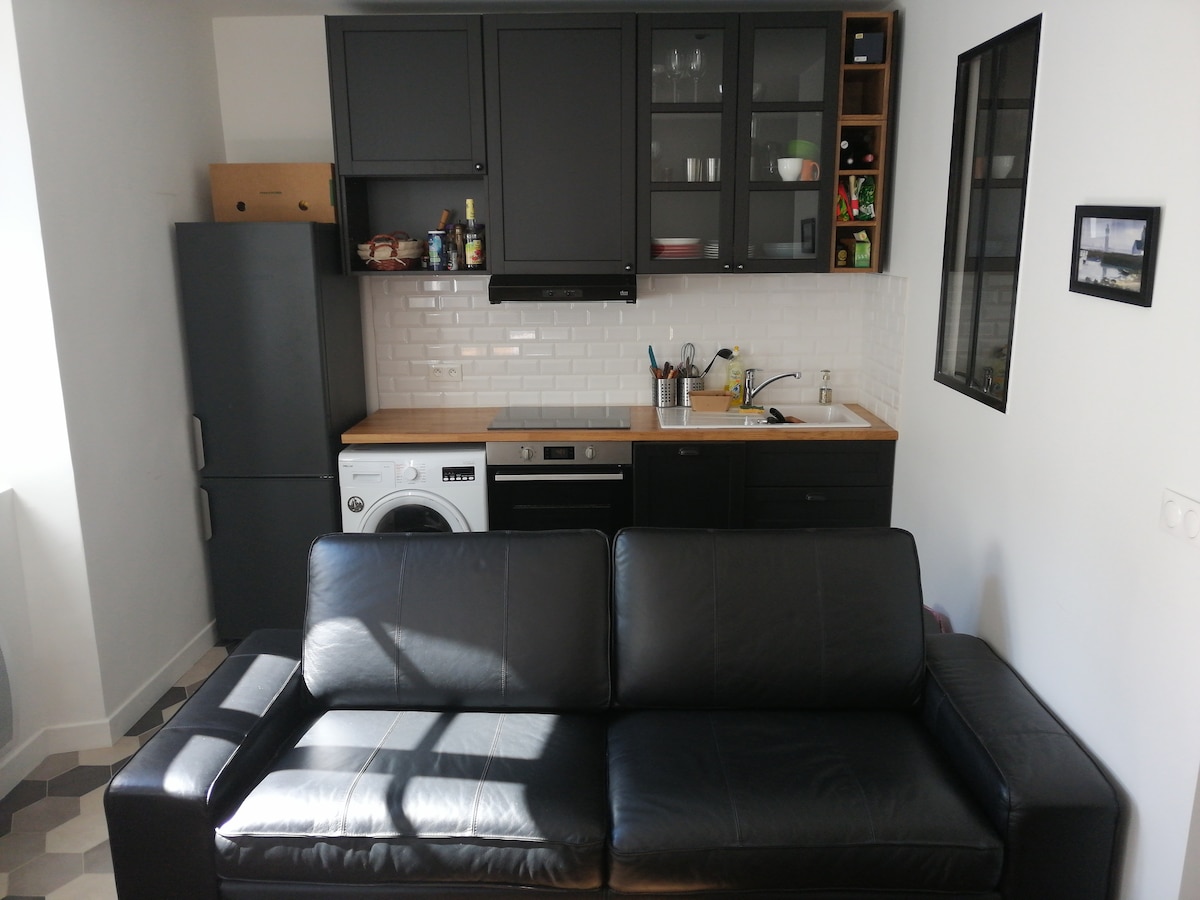 A modern kitchen area is displayed, featuring sleek black cabinetry and a contrasting light backsplash. Kitchen appliances, including a refrigerator, washing machine, and dishwasher, are arranged for accessibility. Natural light enters through a small window, highlighting the wooden countertop and the organized use of space.